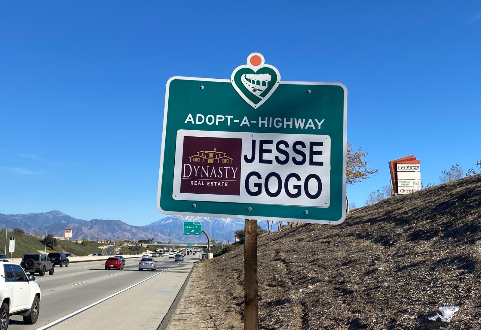 Jesse Gogo Adopt-A-Highway sign above the SR-210 freeway near the Ayala Drive exit, with snow-capped mountains behind.