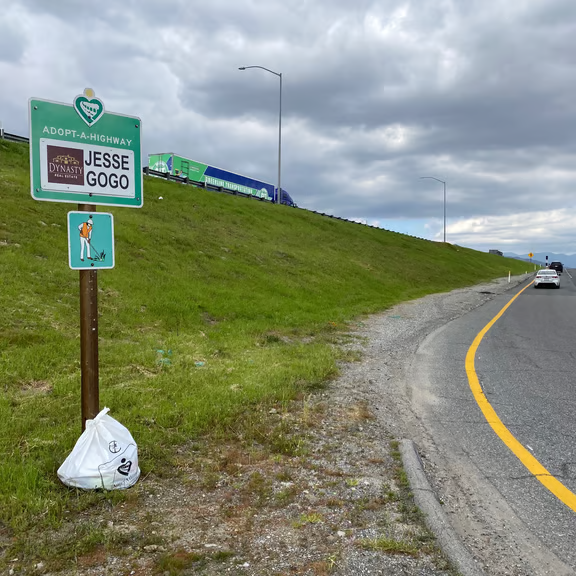 Jesse Gogo Adopt-A-Highway sign on a grassy on-ramp slope with a filled white cleanup bag at the base of the post under cloudy skies.
